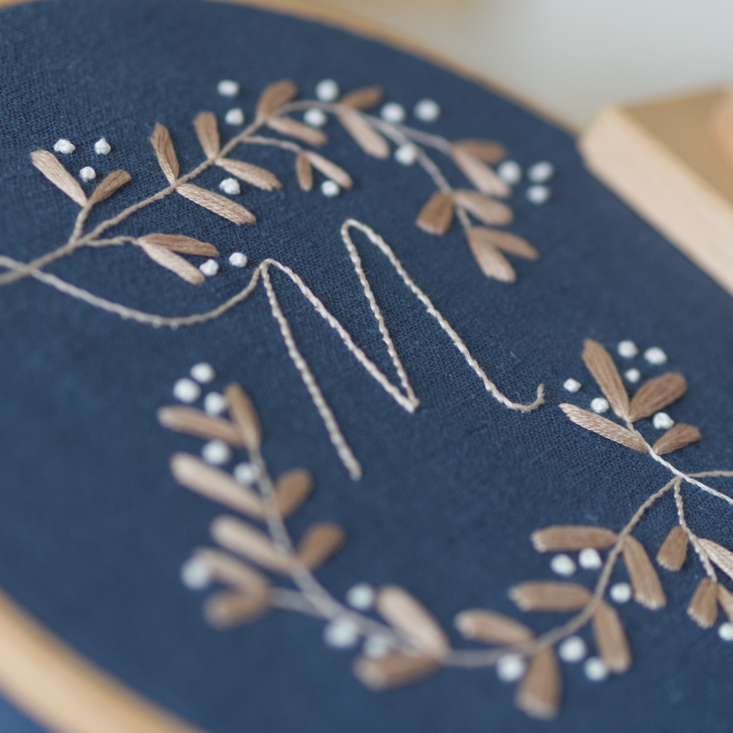 Close up of a golden beige botanical monogram embroidery design with the letter "M" on a dark blue fabric in an embroidery hoop.