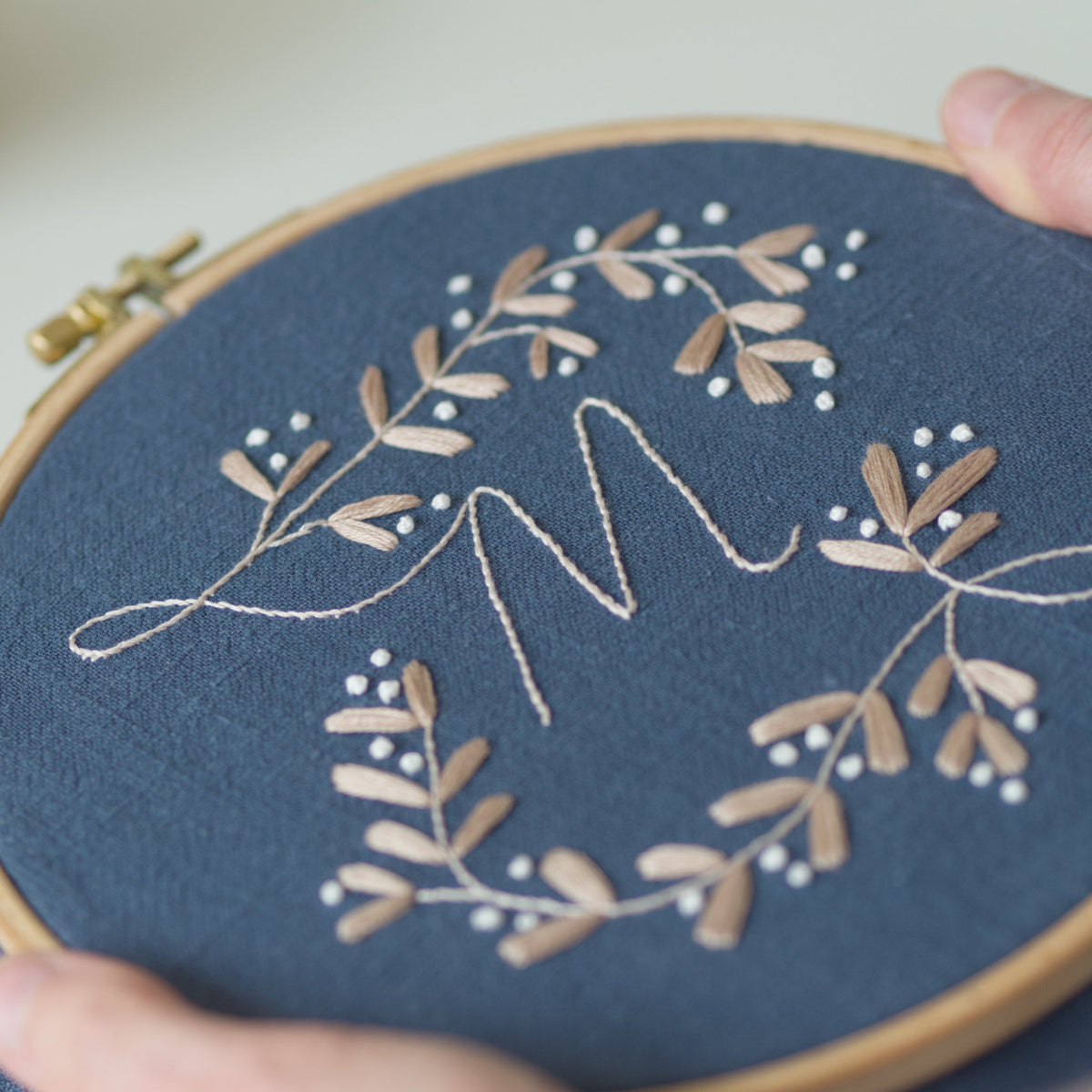 A close-up of a hand holding a wooden embroidery hoop with deep blue fabric. The embroidery features a light beige monogram letter 'M' surrounded by delicate mistletoe design stitched with beige leaves and small white berries.