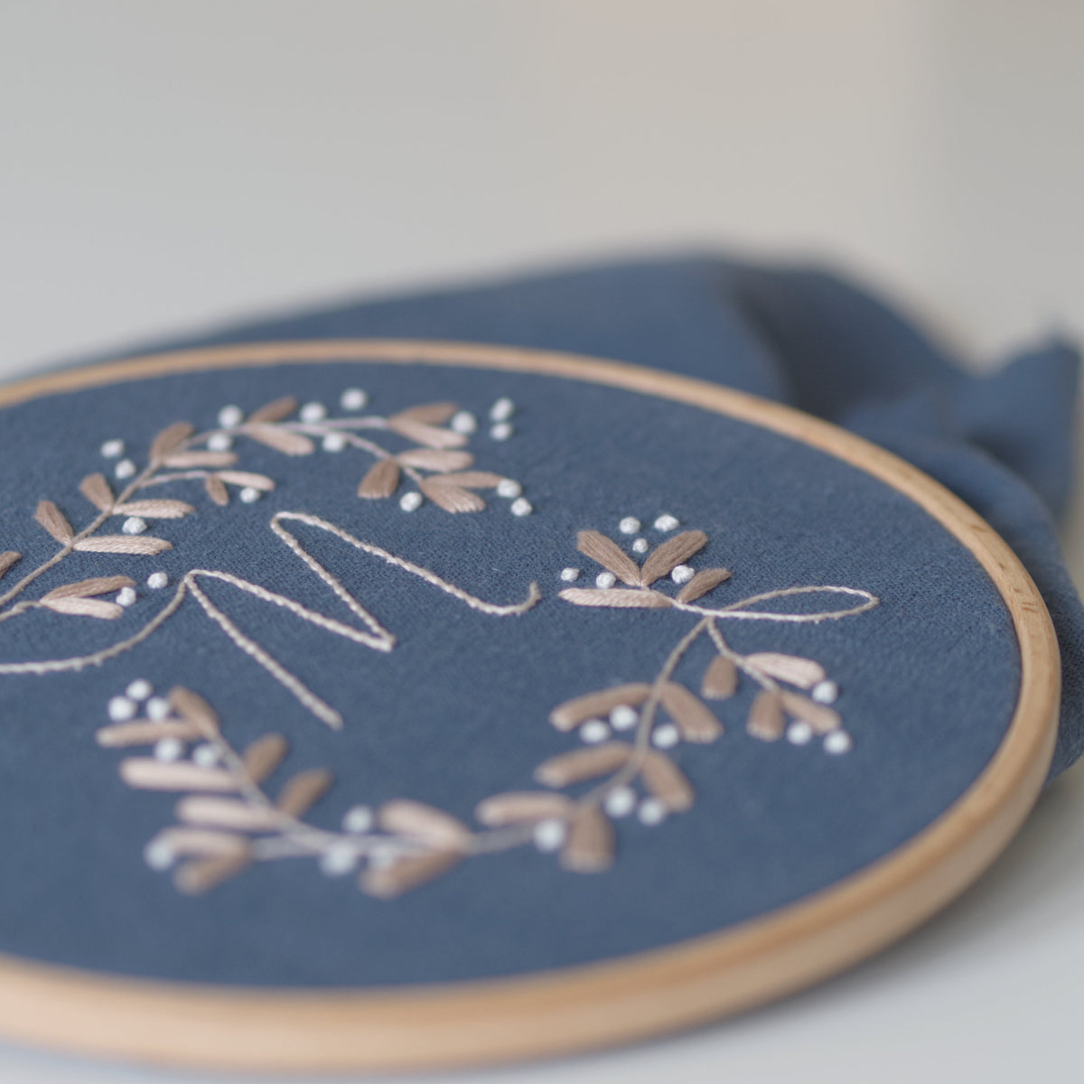 A close-up of a wooden embroidery hoop holding dusty blue fabric with a floral mistletoe wreath design and a monogram in white and beige thread.