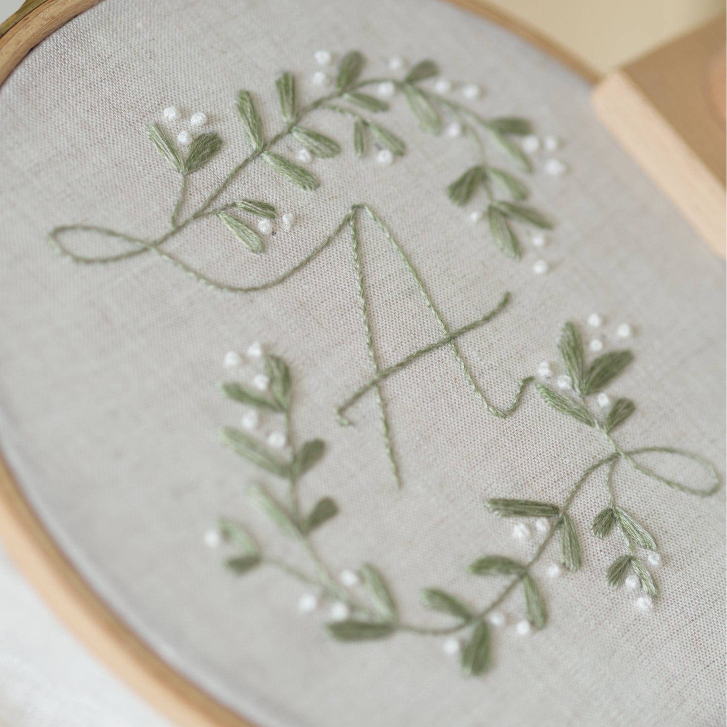 Close-up of a floral mistletoe embroidered monogram design in sage green and white thread on a light beige fabric, held in a wooden embroidery hoop.