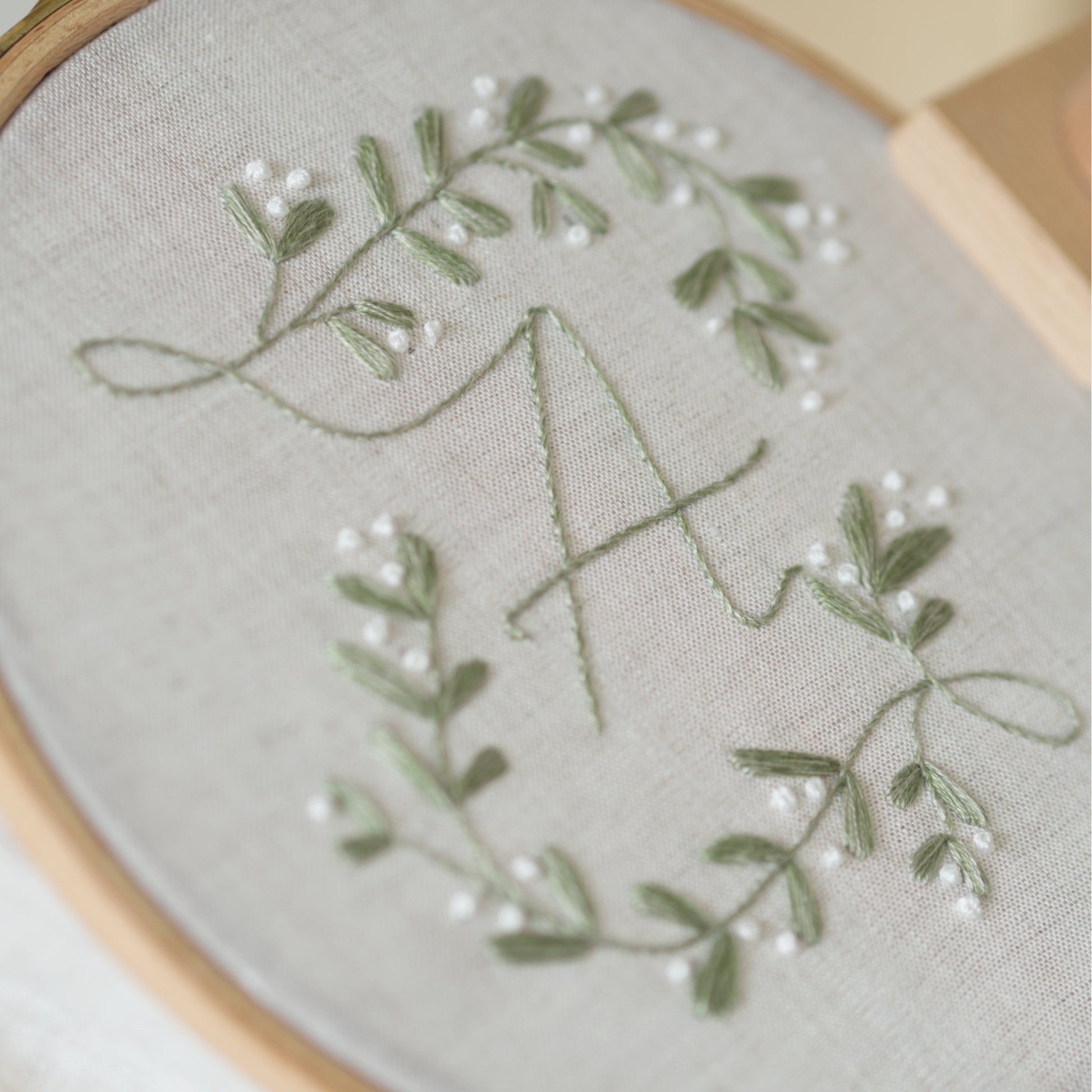 Close-up of a floral mistletoe embroidered monogram design in sage green and white thread on a light beige fabric, held in a wooden embroidery hoop.