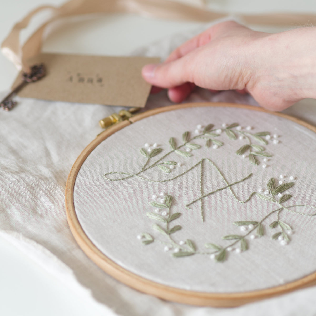 A close-up of a hand holding a kraft paper tag next to a wooden embroidery hoop containing a light green mistletoe floral wreath and a monogram letter 'A' stitched on beige linen fabric.