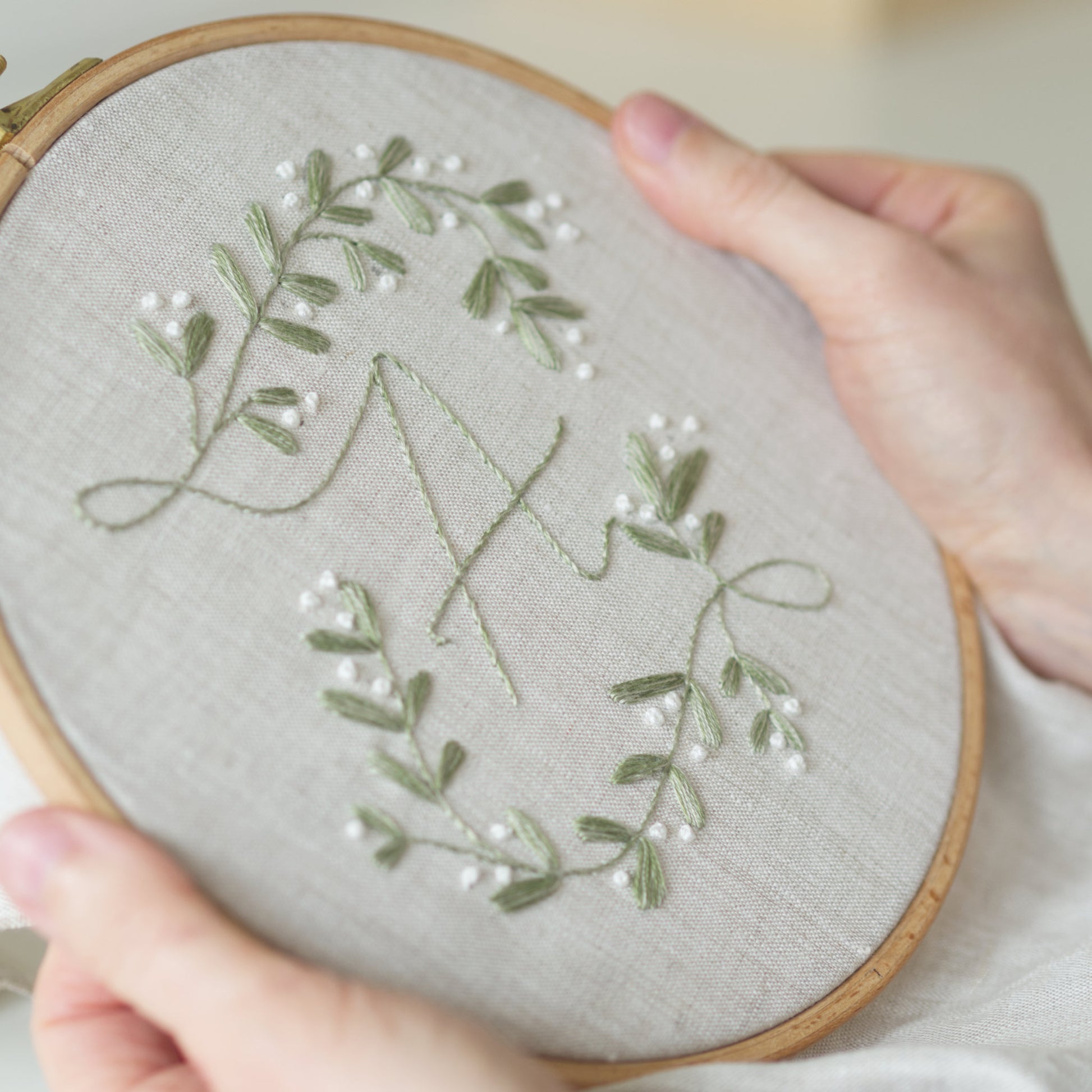 A close-up of hands holding a wooden embroidery hoop with a light beige linen fabric. The embroidery features a sage green monogram letter 'M' surrounded by delicate mistletoe with green leafy vines and small white pearl-like beads.