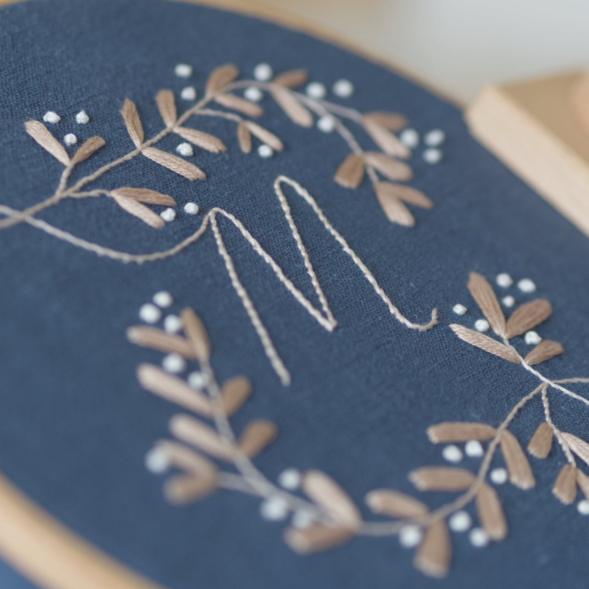 Close-up of a deep blue fabric monogram embroidery hoop, featuring a stitched letter 'M' surrounded by delicate beige and white mistletoe with berries and leafy branches.