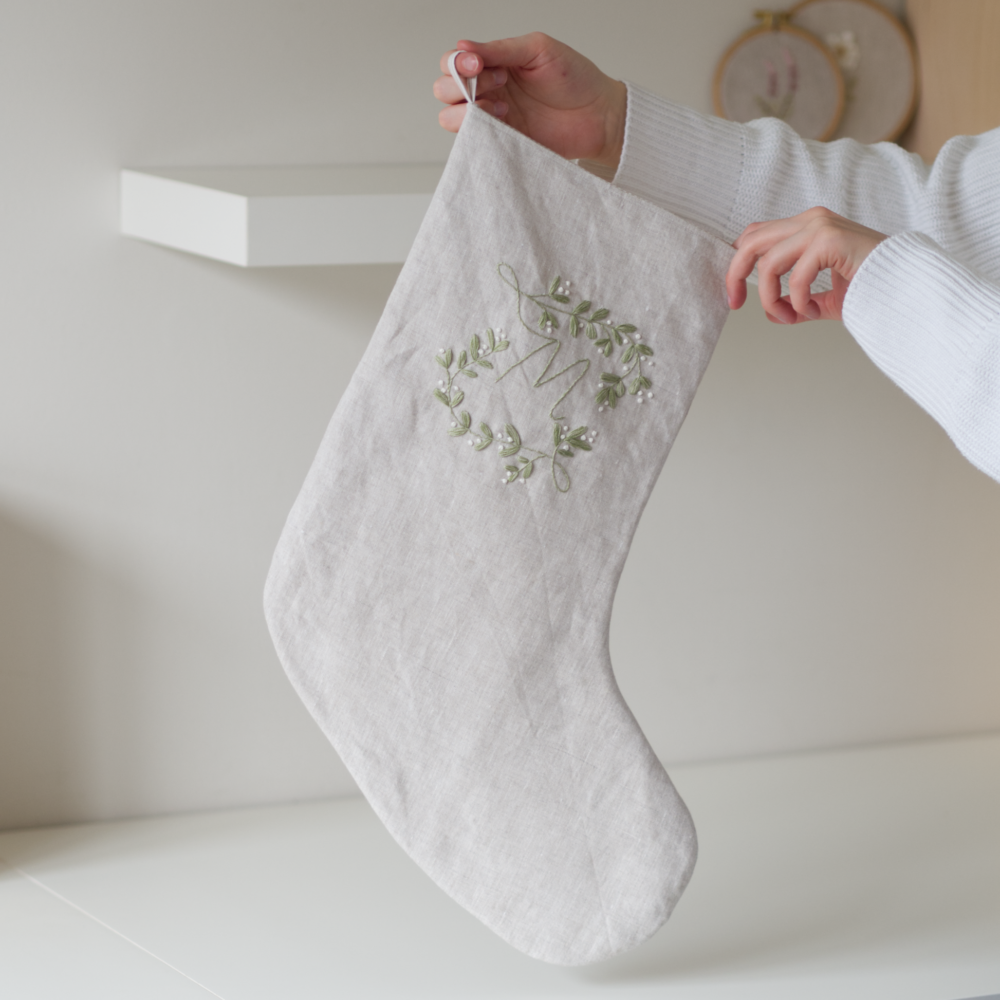 Person holding a stocking in beige linen and embellished with a botanical monogram embroidery design with the letter "M" stitched in green and white thread.