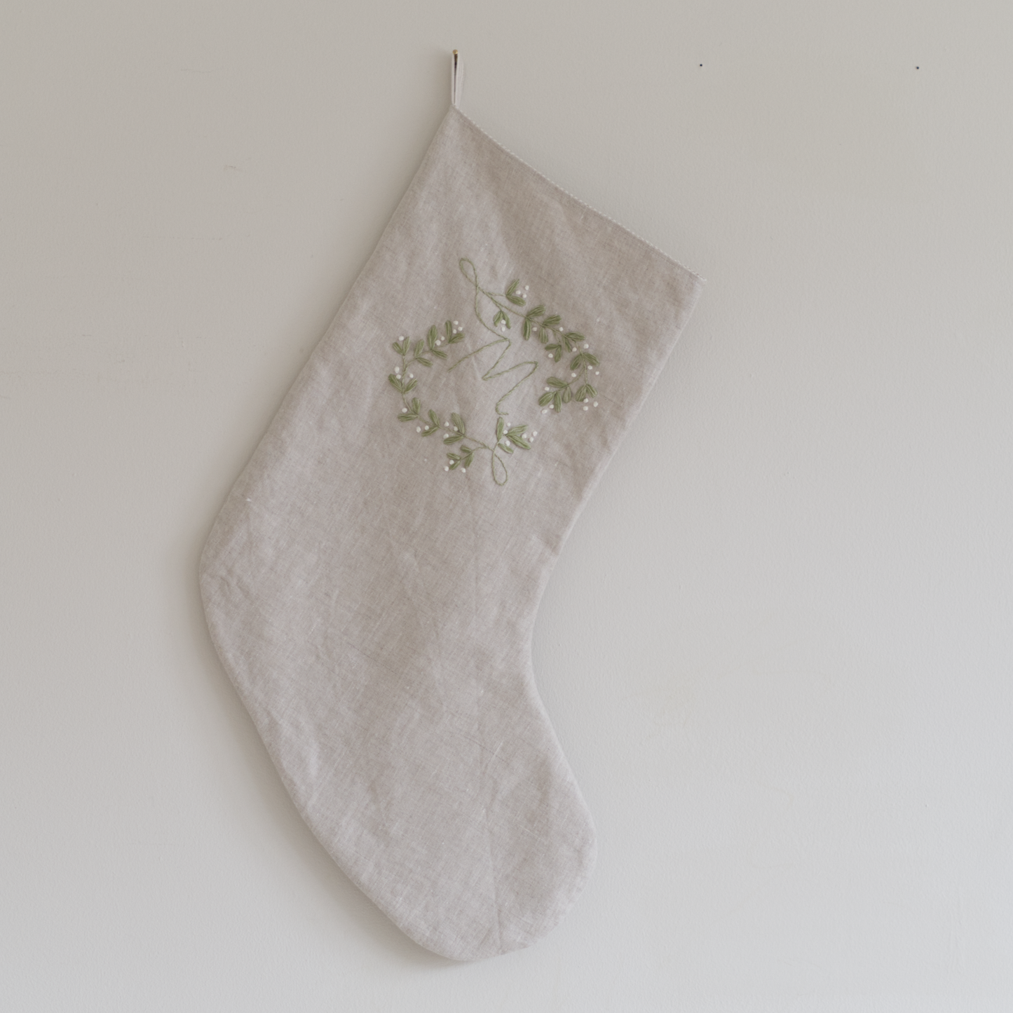 Stocking in beige linen and embellished with a botanical monogram embroidery design with the letter "M" stitched in green and white thread, hung on a wall.