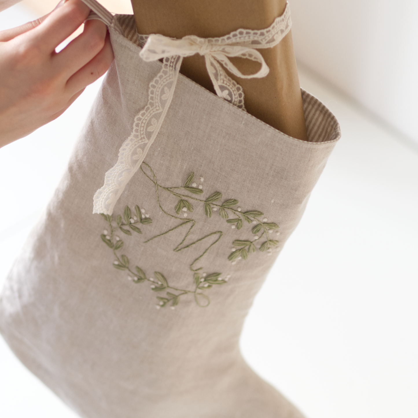 Person putting a brown paper package tied up in cream lace into a beige linen stocking with a botanical embroidery design of the letter "M" stitched in muted green and white.