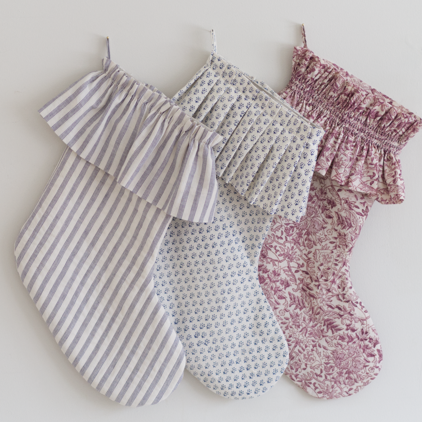 Three hand sewn stockings with varying skirts of gathers, pleats, or bead smocking, and fabrics of blue stripes, block print and pink floral.