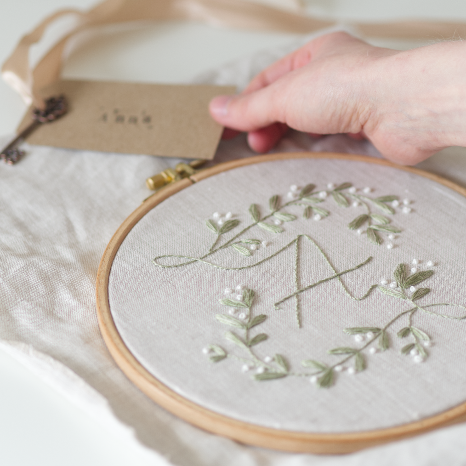 Close up of a botanical monogram embroidery design with the letter "A" in muted green and white thread on a beige linen fabric. A person is setting a brown gift tag with bronze key, gold ribbon and the name "Anna" stamped on it.