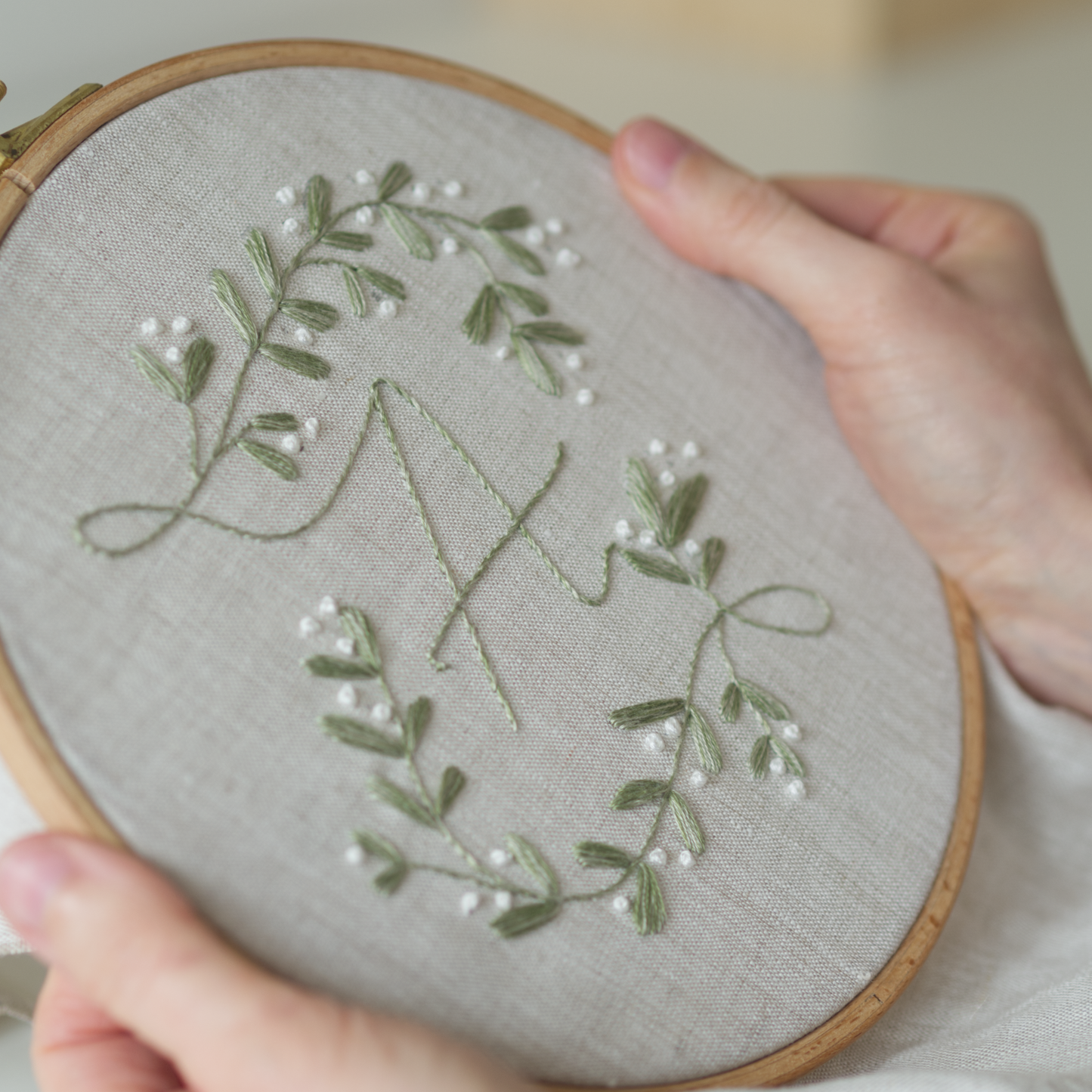 Embroidery hoop with green leaves and white flowers on grey fabric held by a person.