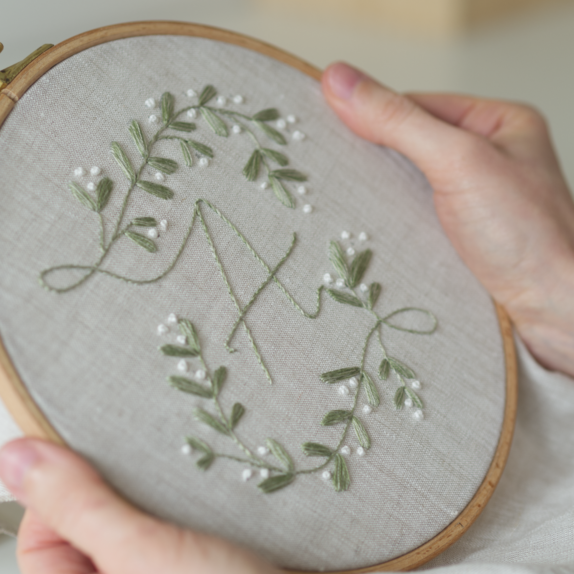 Embroidery hoop with green leaves and white flowers on grey fabric held by a person.
