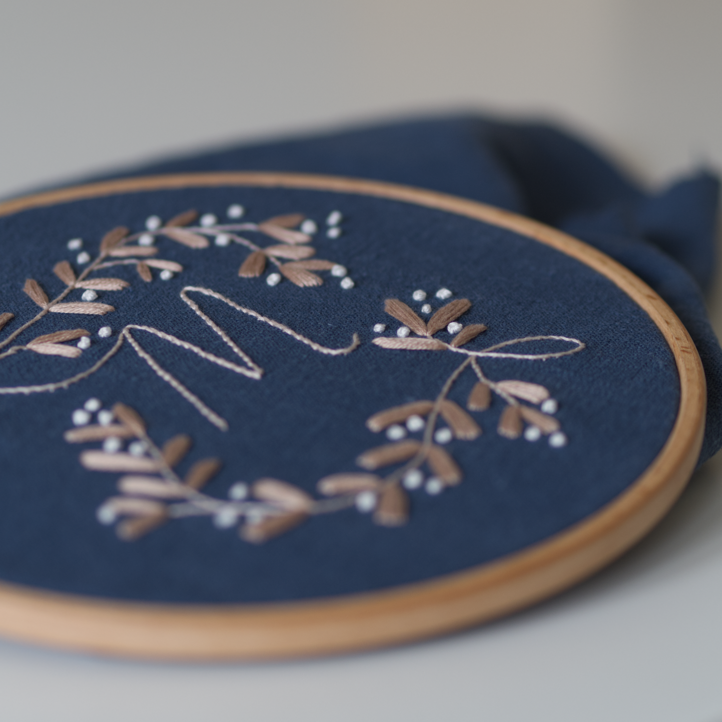 Close up of a golden beige botanical monogram embroidery design with the letter "M" on a dark blue fabric in an embroidery hoop. 