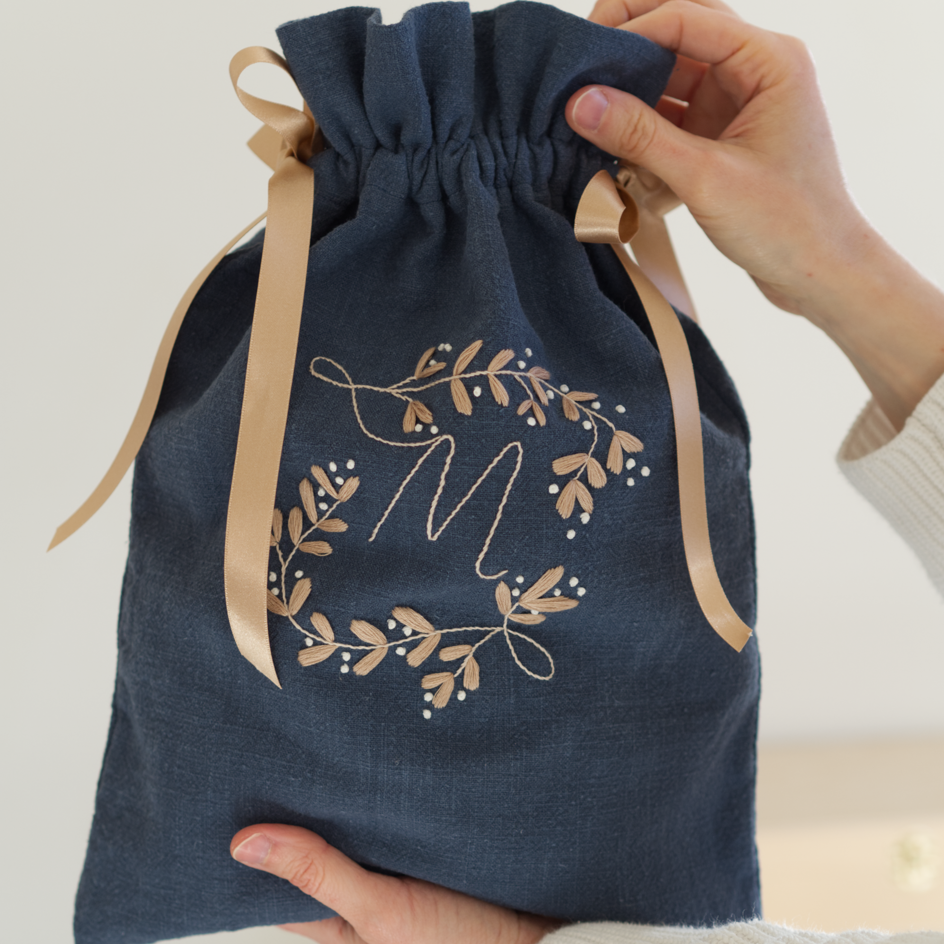 Person holding a drawstring bag in dark blue linen fabric with gold and white botanical monogram embroidery design with the letter "M".
