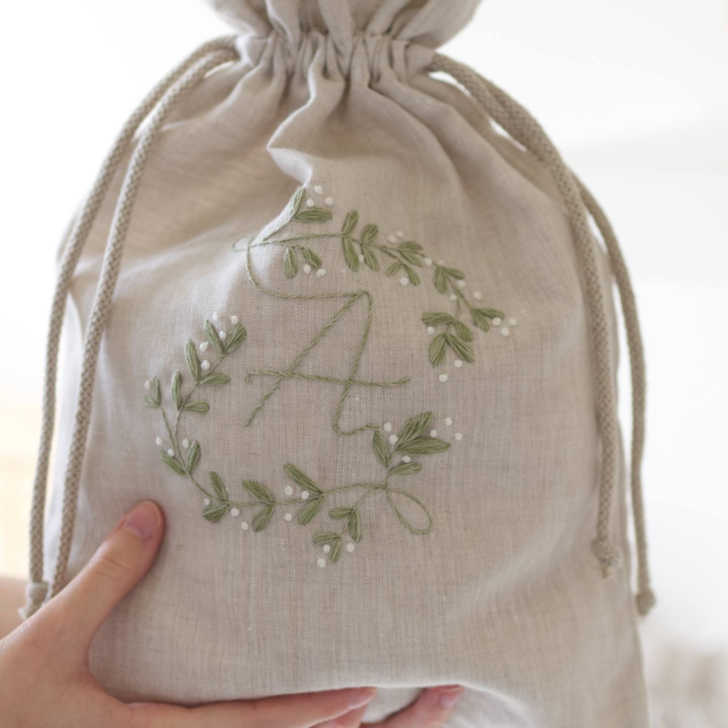 Embroidered design in green thread on a linen beige drawstring bag which a person is holding.