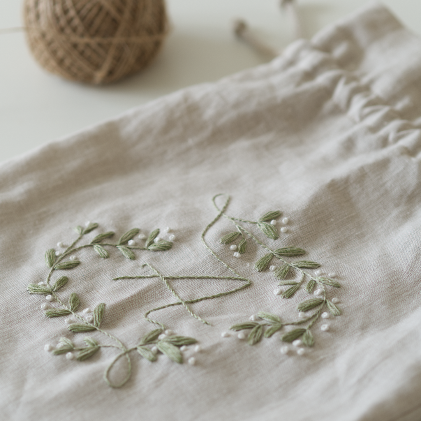 Botanical embroidery design in green thread on a linen beige drawstring bag with a ball of twine in the background