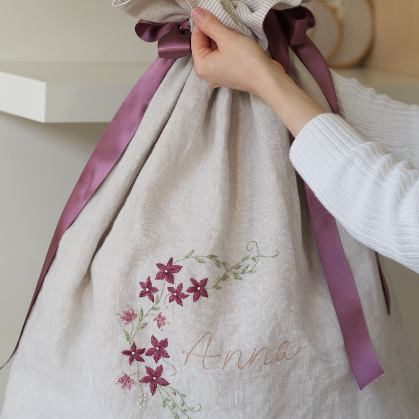 Person holding a festive sack with purple drawstring ribbons. The sack is detailed with a botanical embroidery design of the name "Anna" and a floral vine.