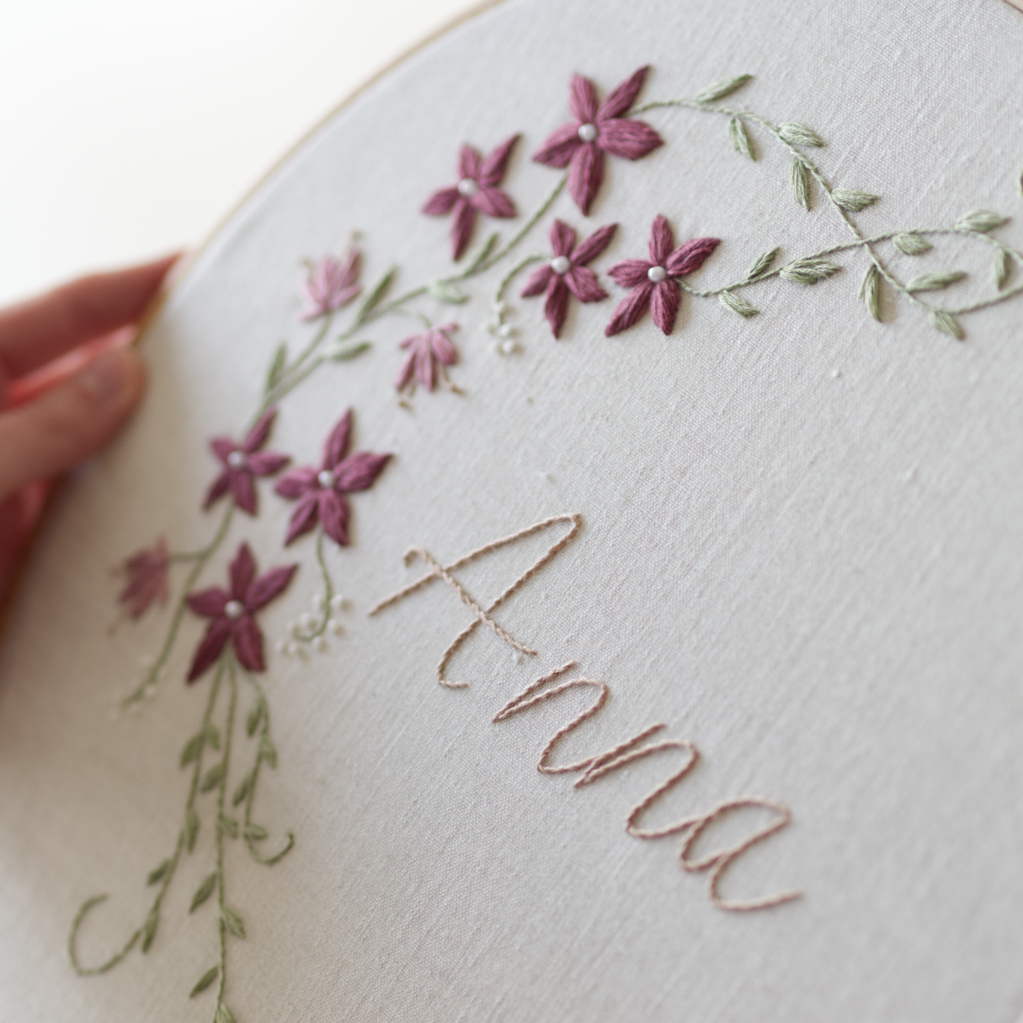 Botanical embroidery design with the name Anna in beige, and a winter vine in green with white berries and plum coloured flowers branching out. 