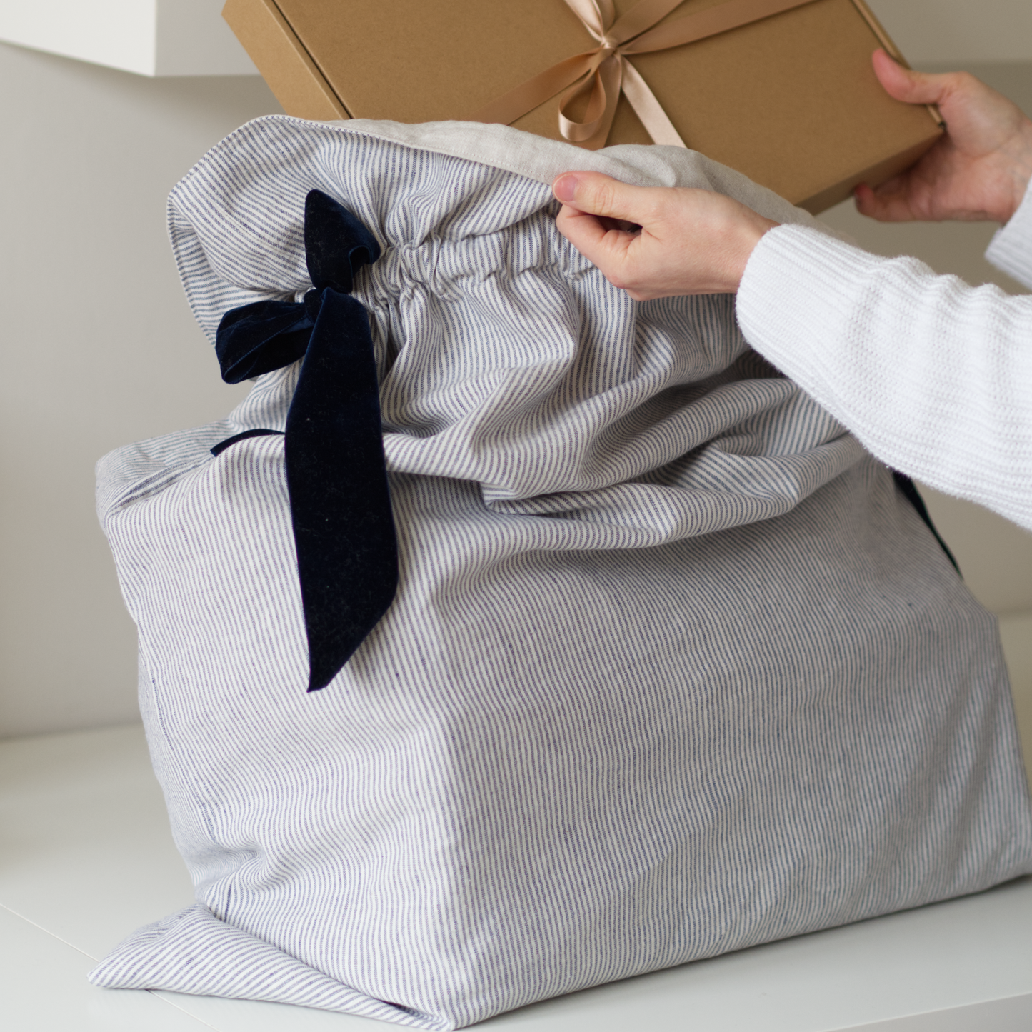Festive sack in striped blue linen fabric with big navy velvet bows. Hands are putting a brown package tied in gold ribbon into it.