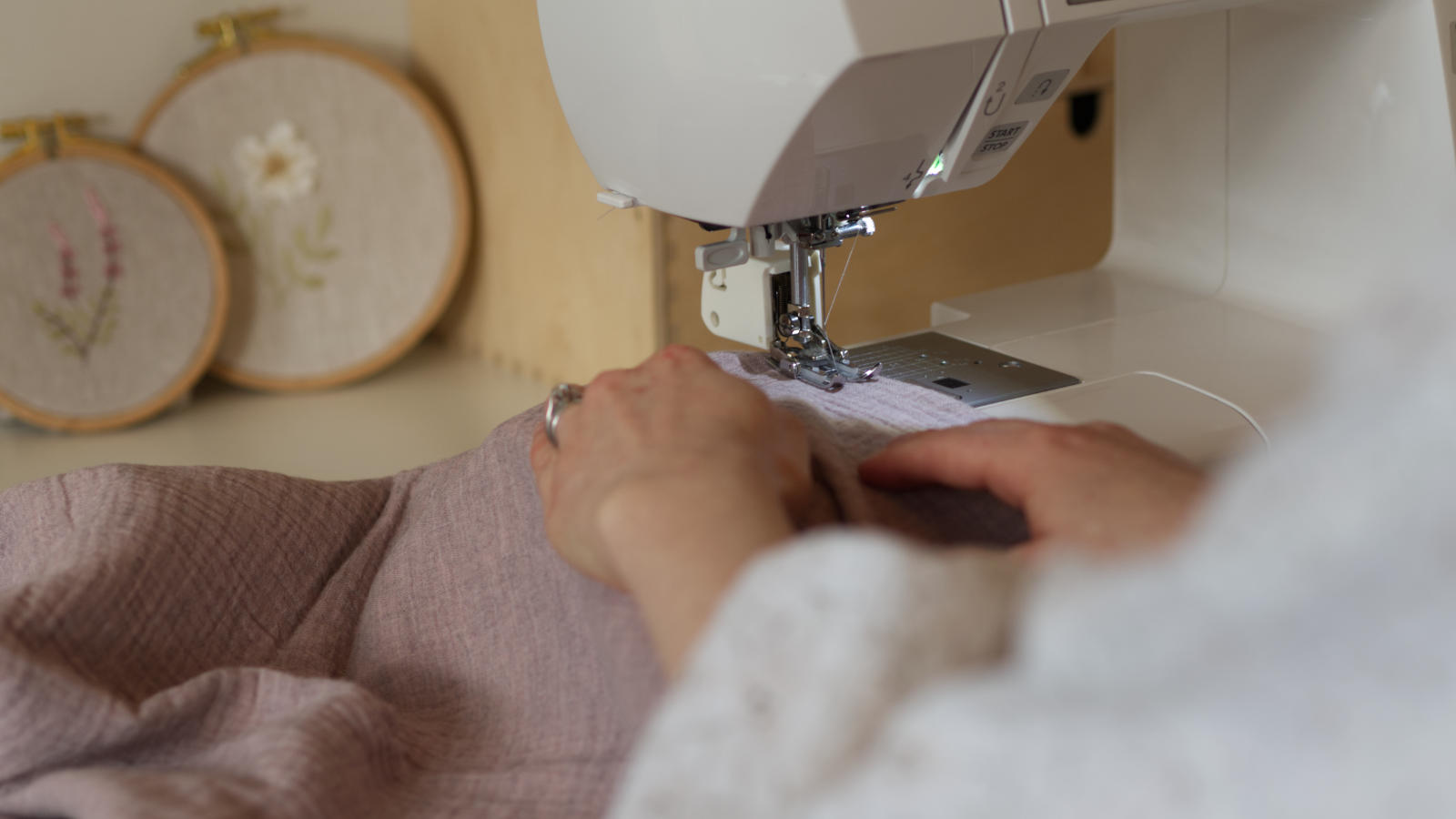 Hands guiding fabric through sewing machine with finished embroidery hoops in the background